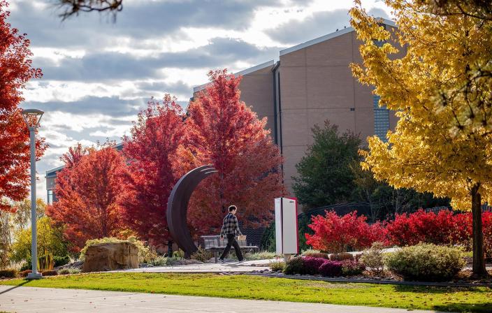 Scenic view of the Greeley Aims Community College campus during fall, showcasing vibrant red and yellow foliage, a modern sculpture, and a student walking along the pathway.