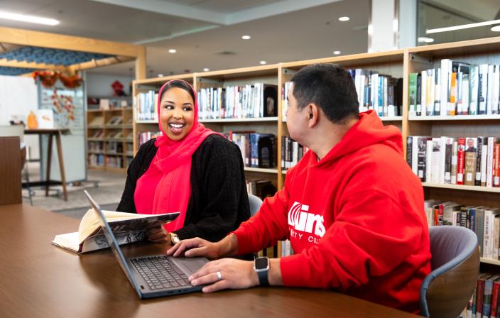 Two students sit at a table amongst the book-stacks. They are smiling at each other