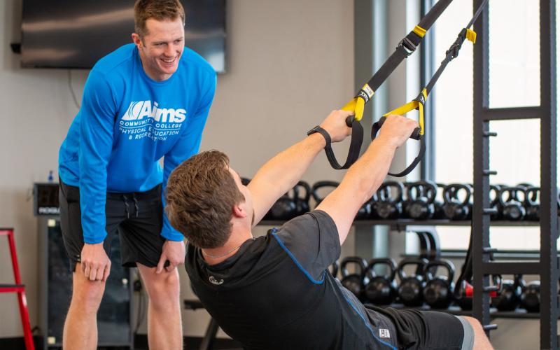 Male trainer encouraging male using weight machine doing overhead row pulls