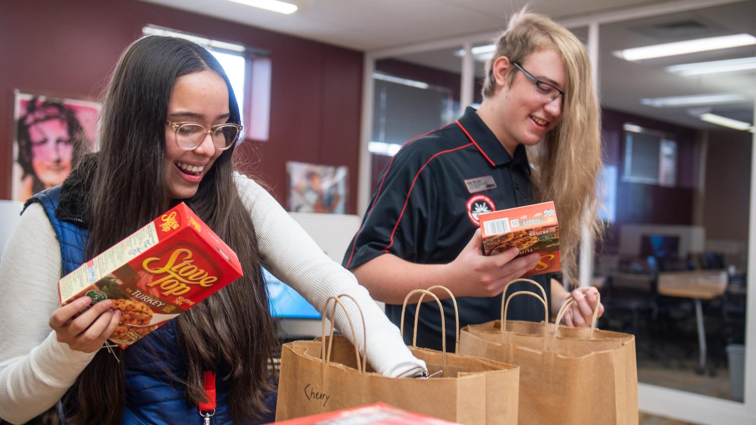 Two smiling Aims students packing groceries into brown paper shopping bags for Thanksgiving meal kits to be distributed through Arty's Pantry