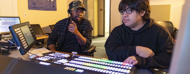 Two students in the Aims Community College Television & Video Production program work together in a control room. One student wears a headset while giving direction, and the other operates a video switcher with rows of brightly lit buttons.